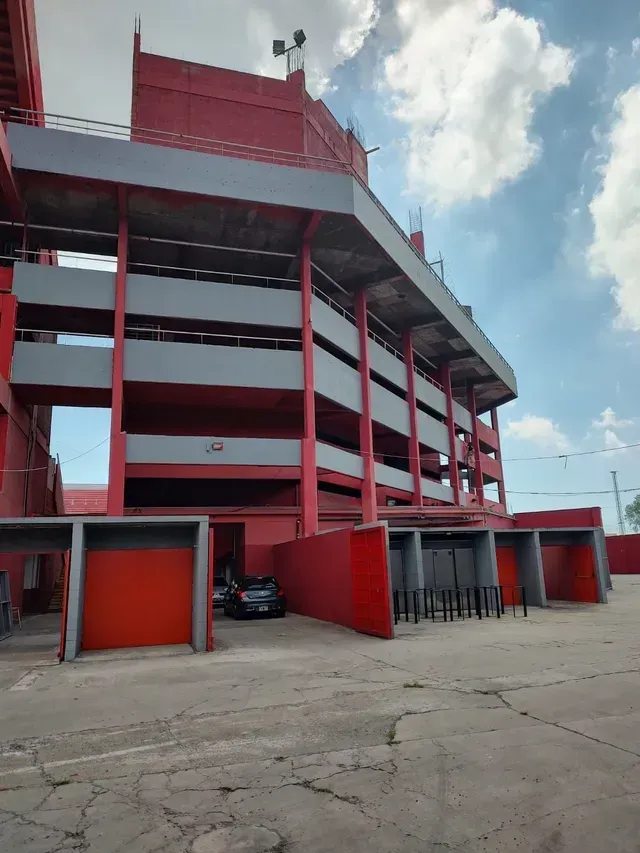 Estadio rojo de varios pisos con niveles abiertos y una explanada de hormigón bajo un cielo nublado.