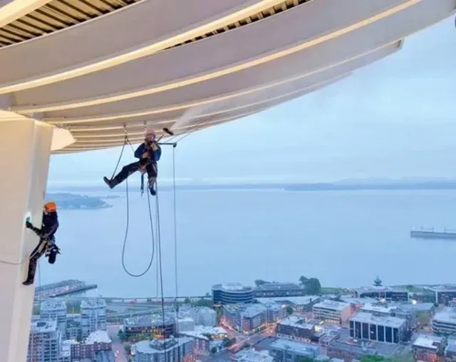 Dos limpiadores de ventanas descienden en rápel desde un rascacielos con vistas al horizonte de una ciudad costera.
