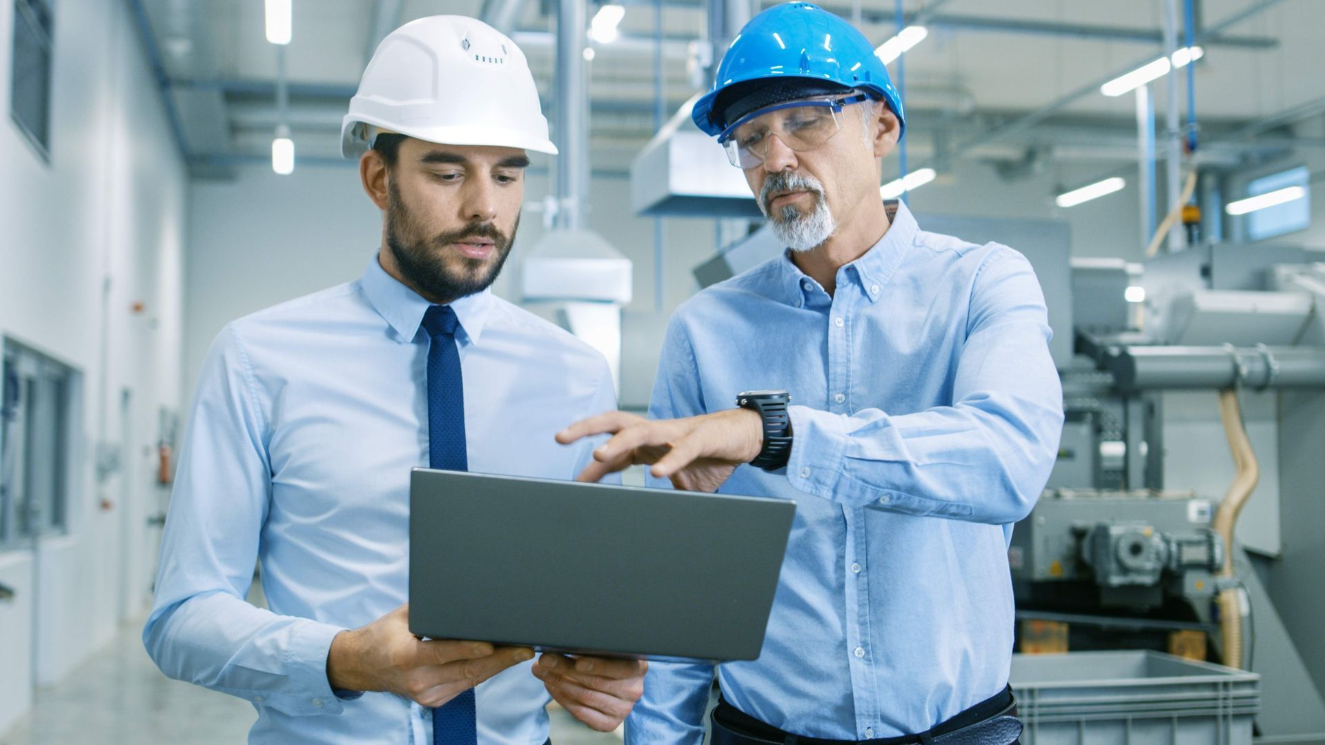 two men in hard hats are looking at a laptop in a factory .