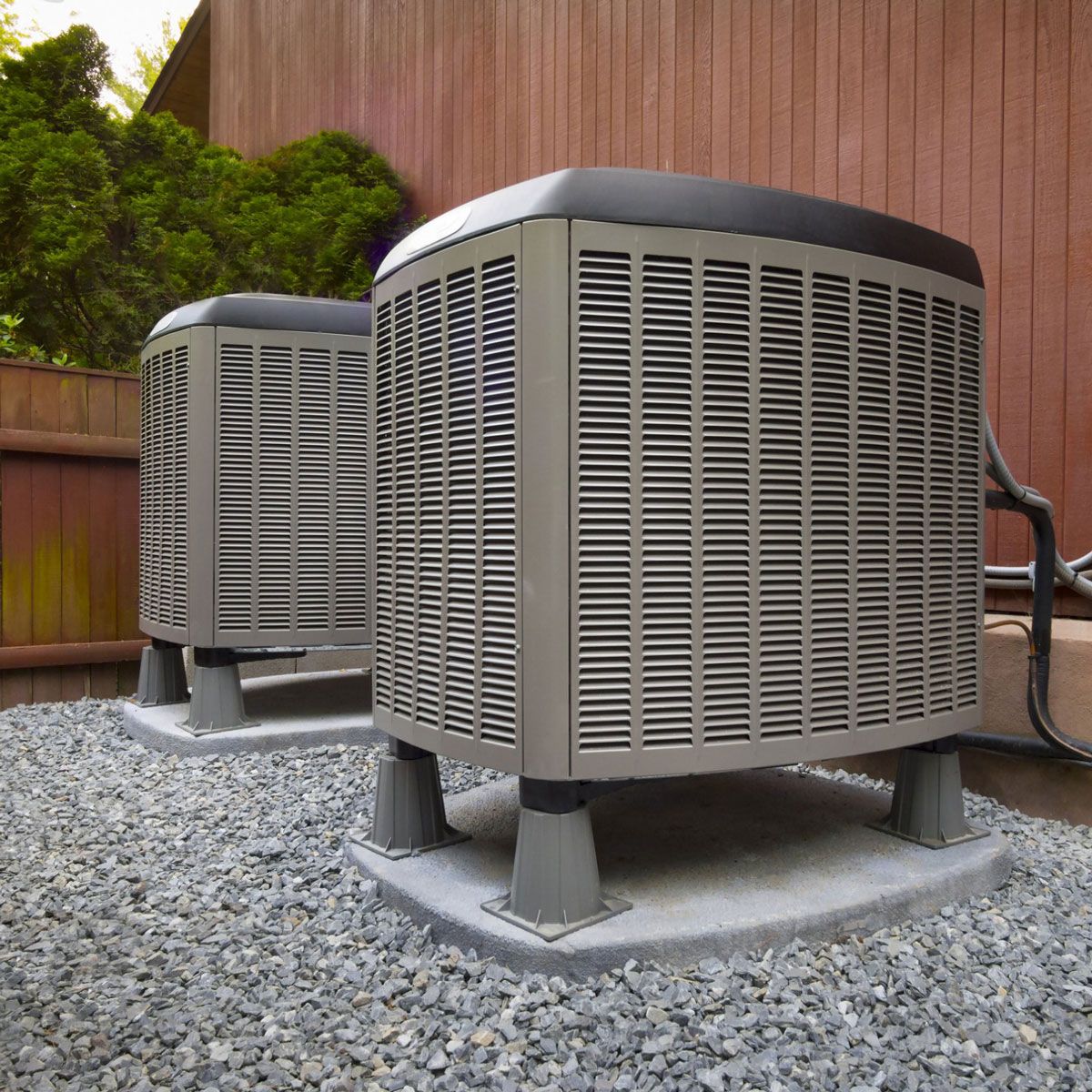 Two air conditioners are sitting on gravel outside of a house. Two air conditioners are sitting on gravel outside of a house.