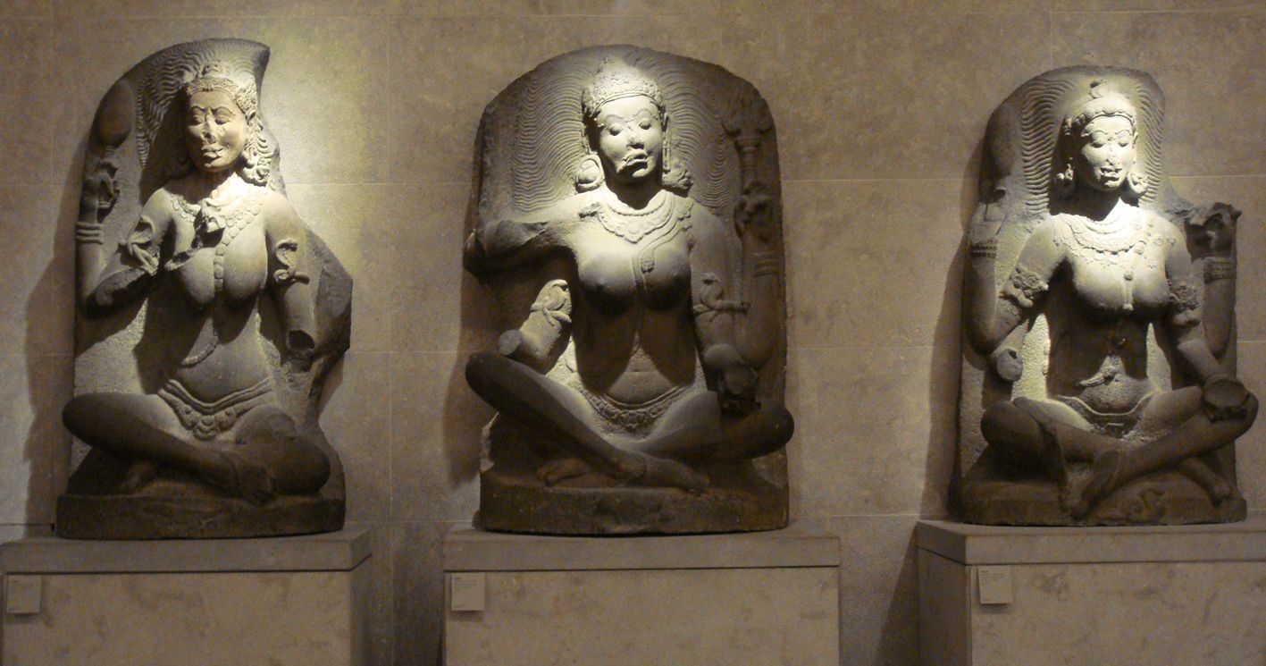 Drie stenen beelden van zittende figuren met sierlijke hoofdtooien en sieraden staan ​​op sokkels tegen een muur.