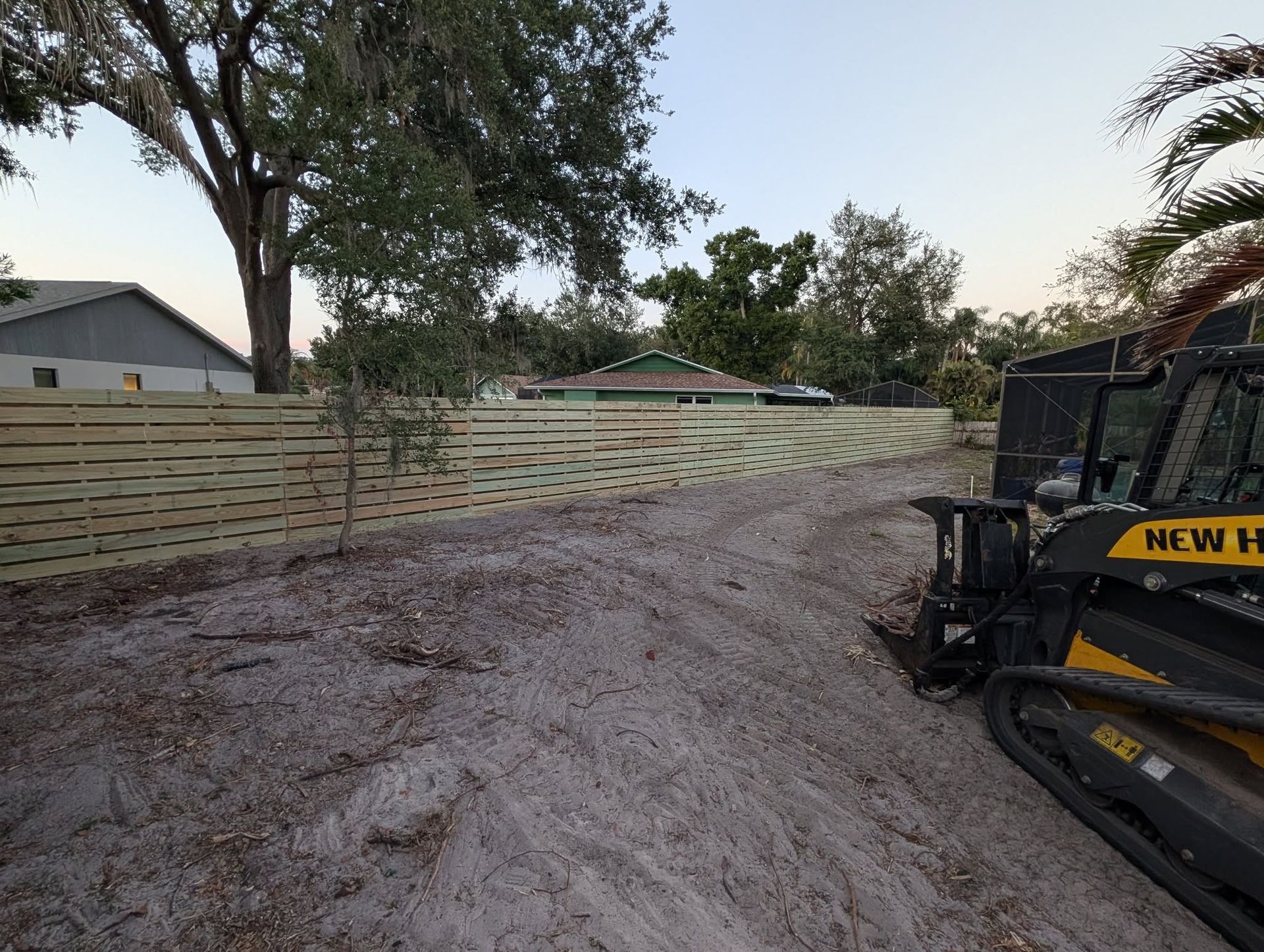 Dirt driveway beside a wooden fence, with a construction machine on the right and trees overhead.