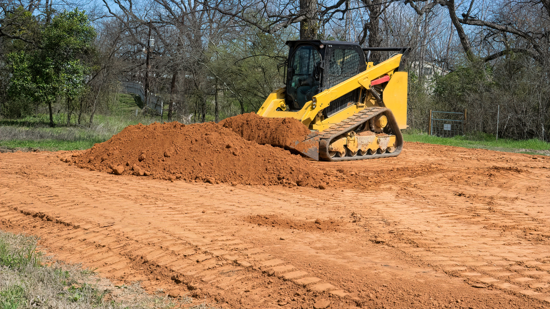 Yellow skid steer pushing a dirt pile on a cleared lot under a tree-lined sky