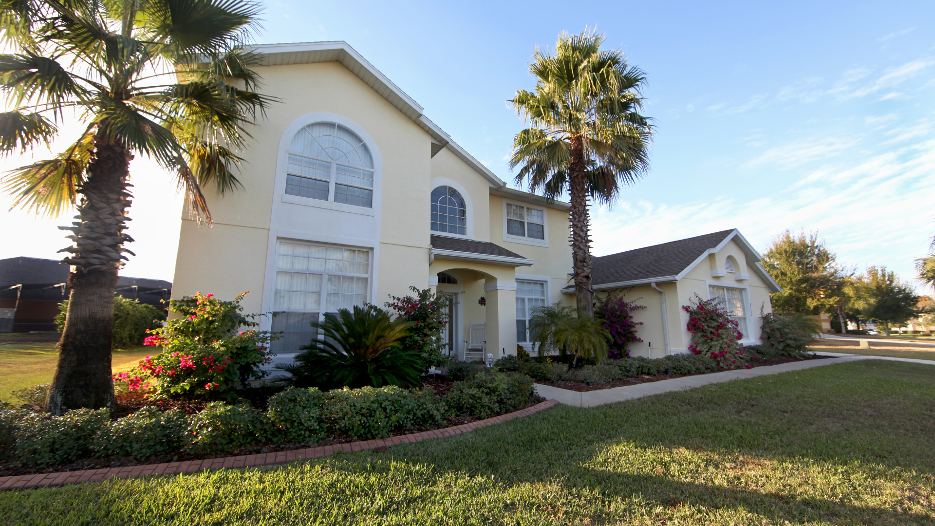White two-story house with arched windows, palm trees, and a landscaped lawn in bright daylight