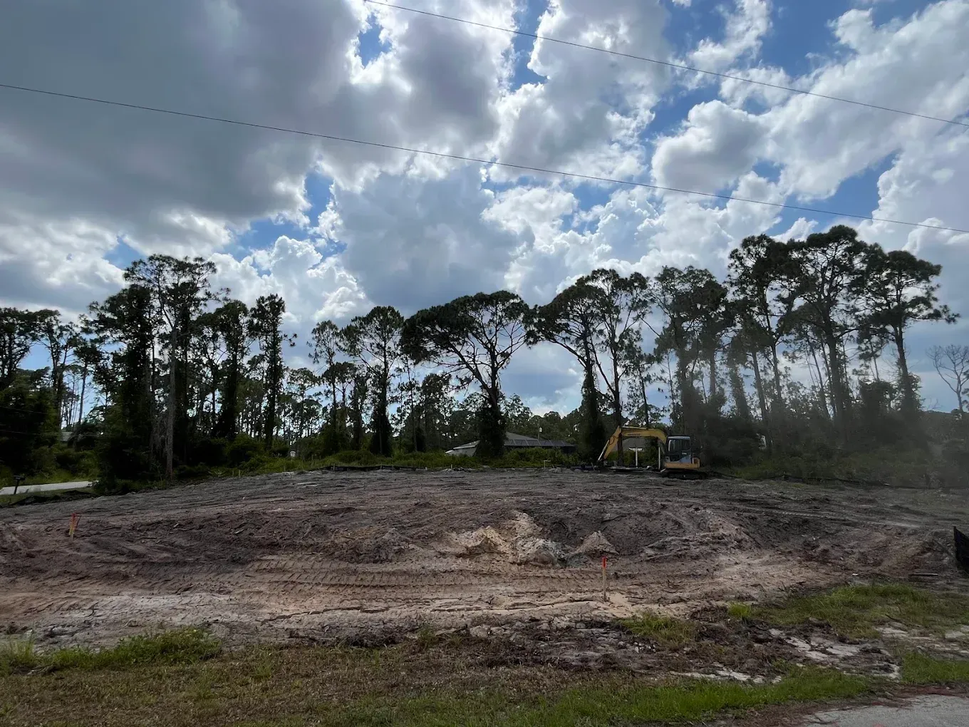 Cleared dirt lot with a small excavator, bordered by trees under a cloudy sky