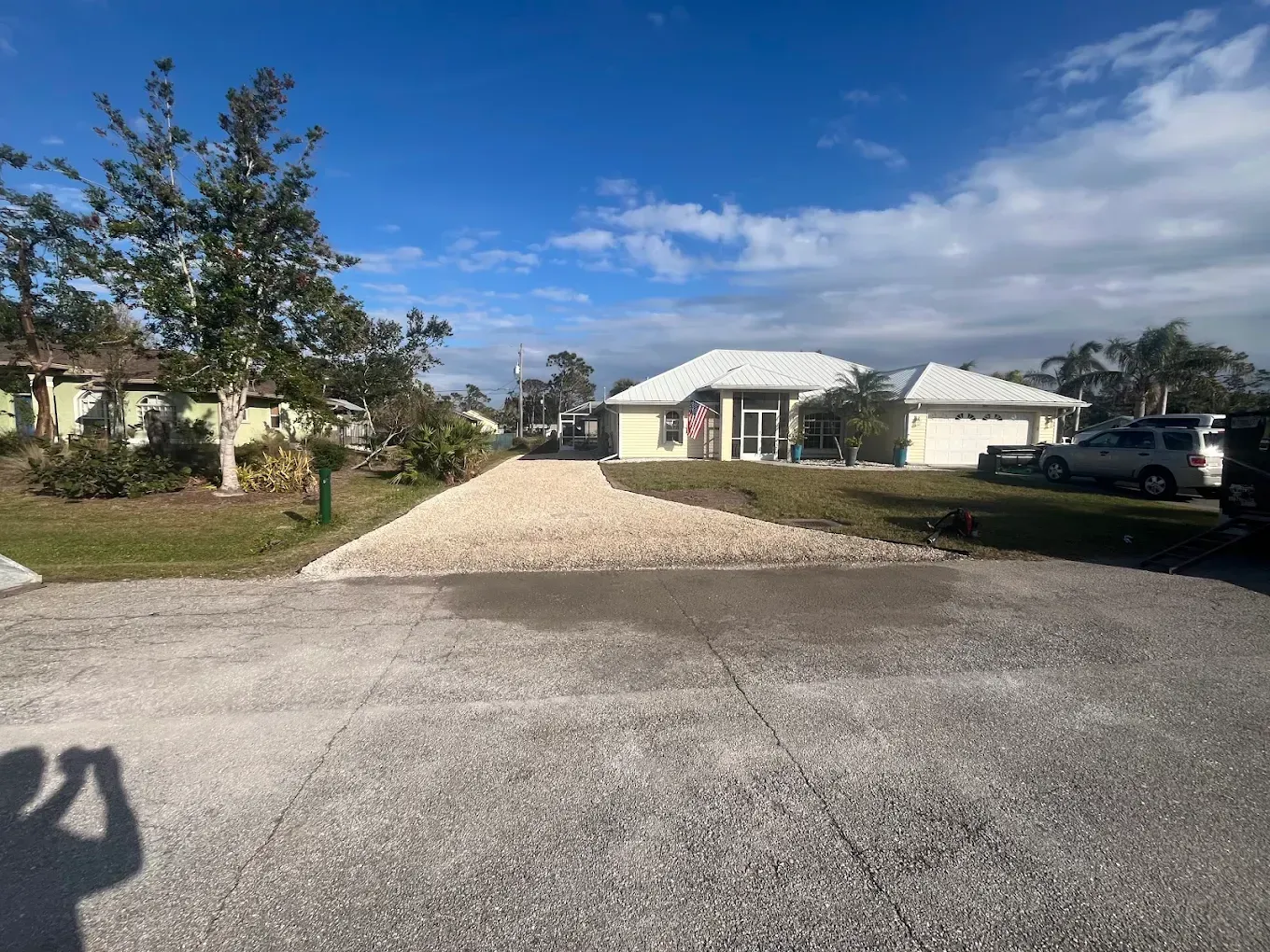 Driveway leading to a white house under a blue sky, with trees and parked vehicles nearby.