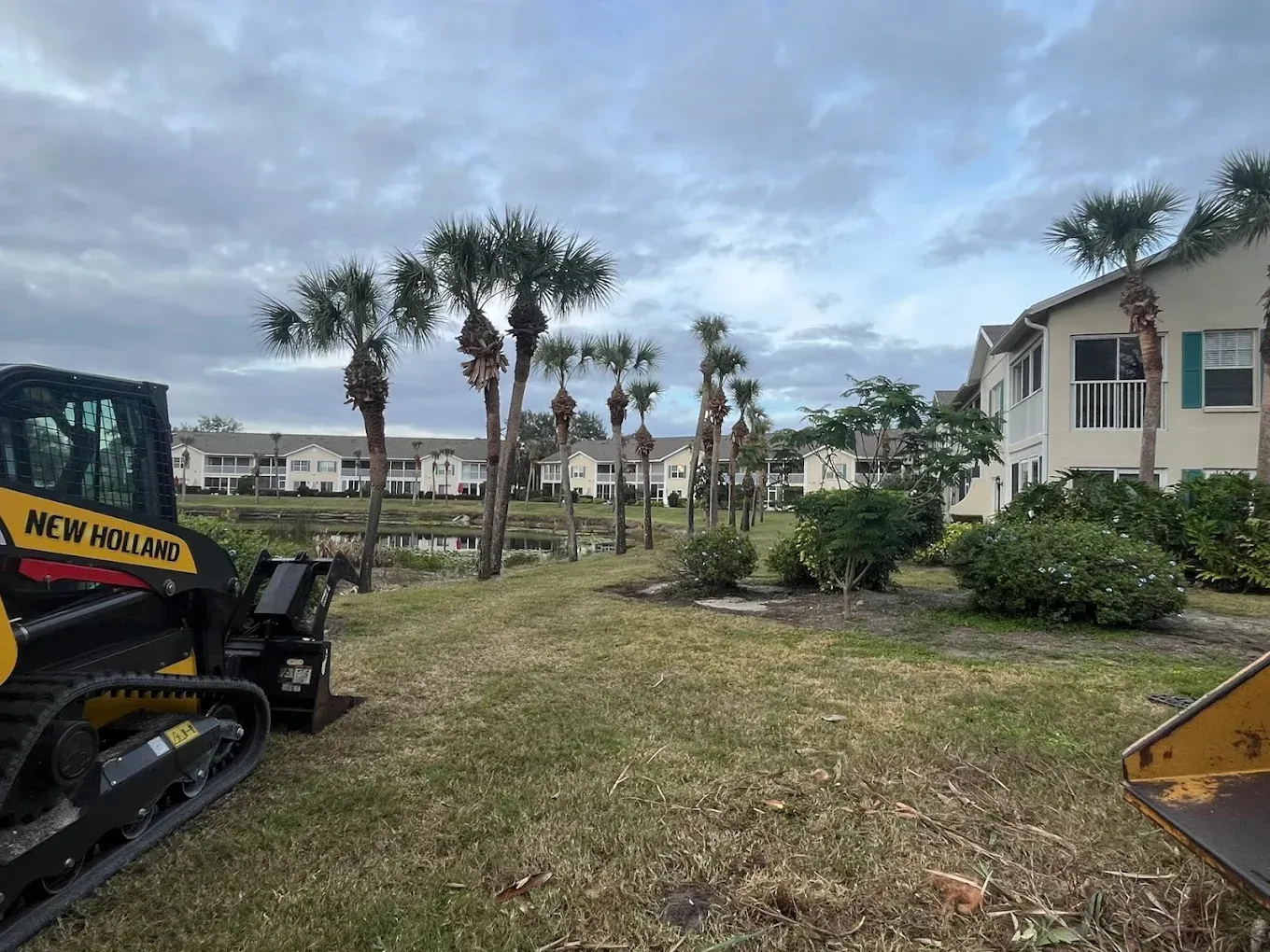 Construction equipment in a grassy lot with palm trees and beachside buildings under a cloudy sky