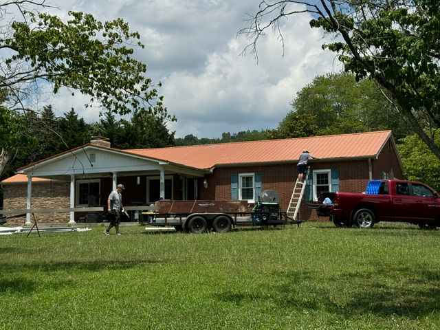 Single-story brick house with orange roof, truck, trailer, ladder, and person in a grassy yard under cloudy sky