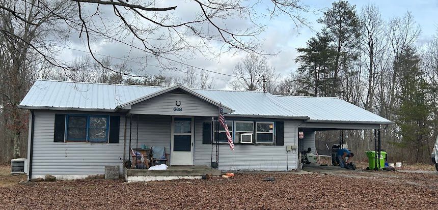 Single-story gray house with a snow-dusted roof, front porch, and trees in the yard