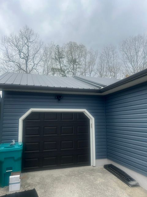 Blue house exterior with a black garage door, trash bin, and snowy yard under an overcast sky