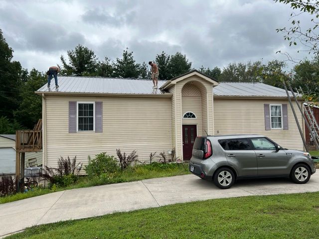 Single-story beige house with silver roof, gray SUV in driveway, and cloudy sky.