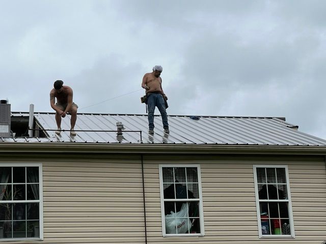 Two workers on a roof, one crouching and one standing near a railing under a cloudy sky