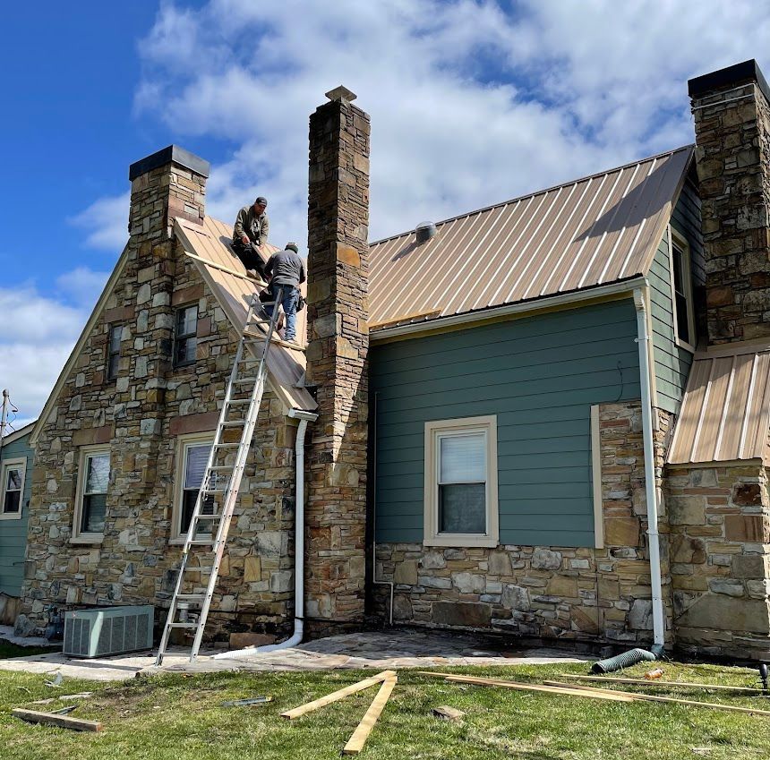 Worker on a ladder repairing the roof of a stone house with blue siding and brick chimneys.