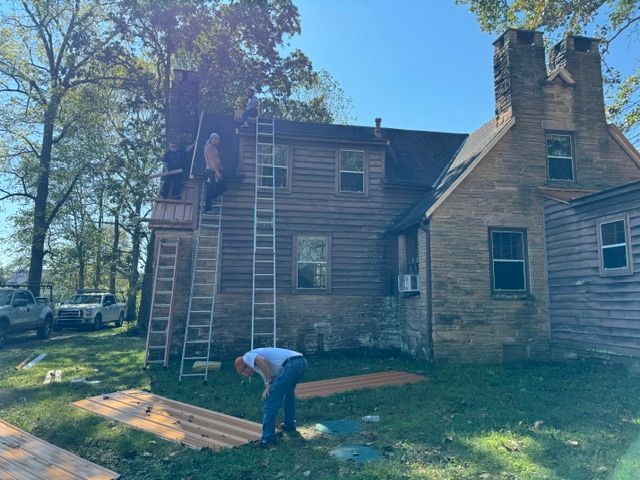Person on a ladder working on the roof of a weathered house while another bends over in the yard.
