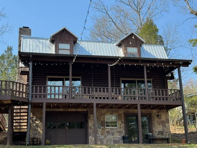 Rustic two-story log cabin with stone base, wooden balcony, and metal roof in a wooded setting