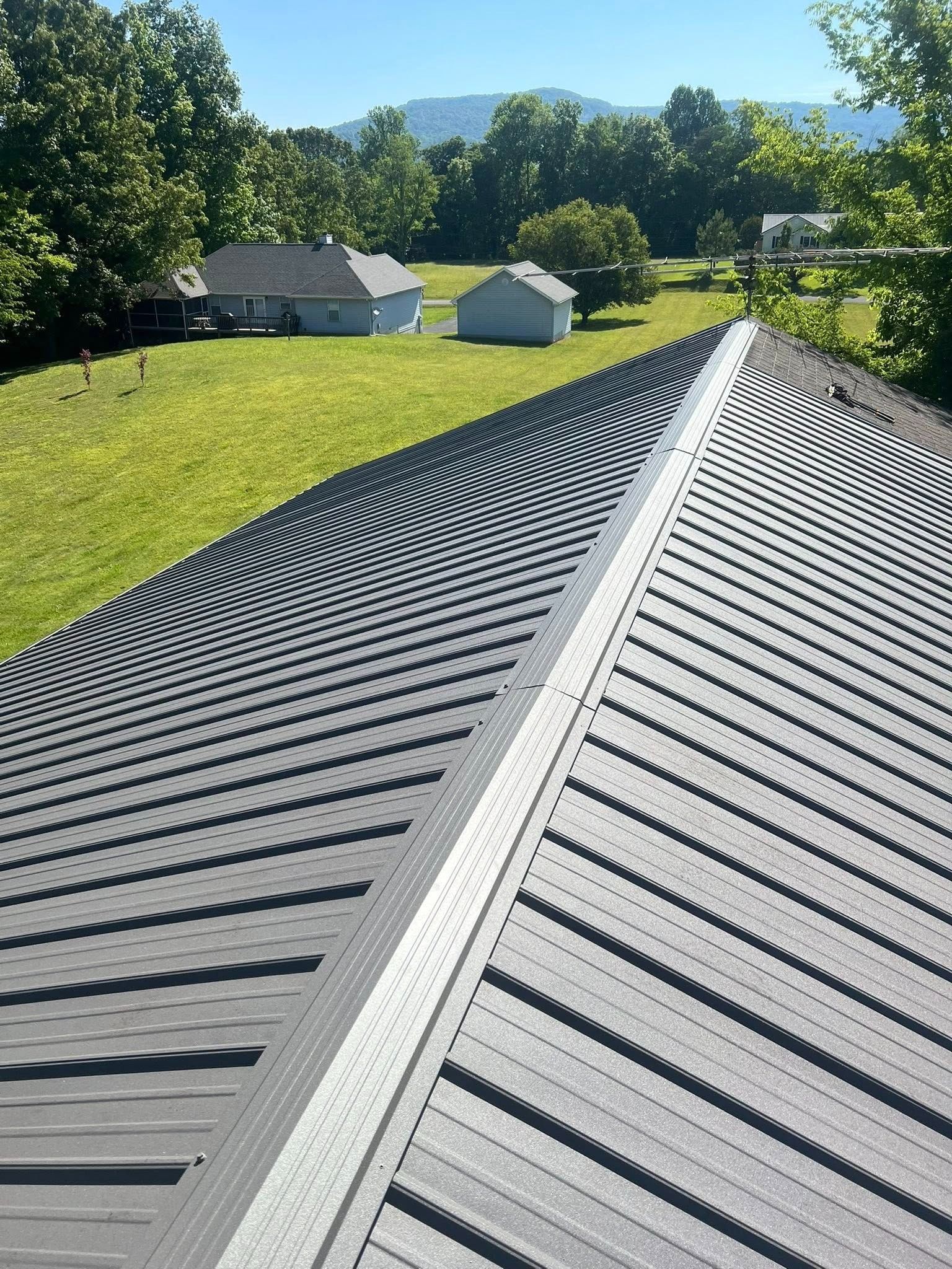 Metal roof ridge with black panels overlooking a grassy yard, houses, and trees in bright sunlight