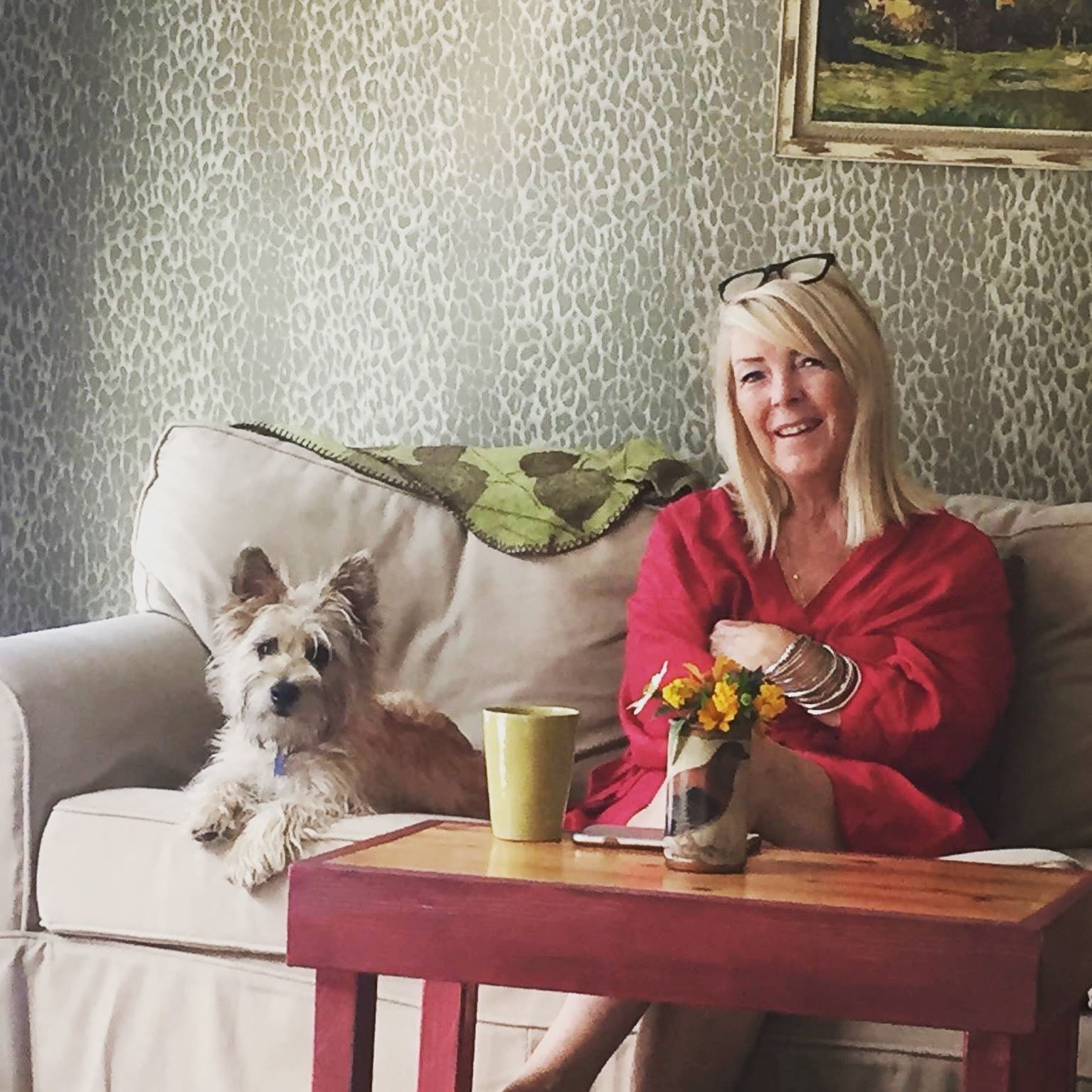 Woman in a red shawl sits on a sofa beside a small dog and a coffee table with flowers and a mug