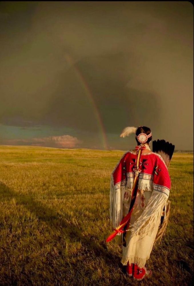 Two people in red and beige outfits stand in a grassy field under a rainbow at dusk