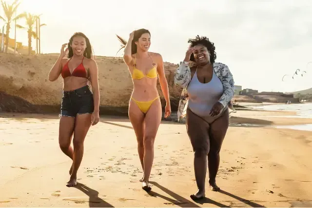 Three women in swimsuits walking together on a sunny beach, smiling near the shoreline.