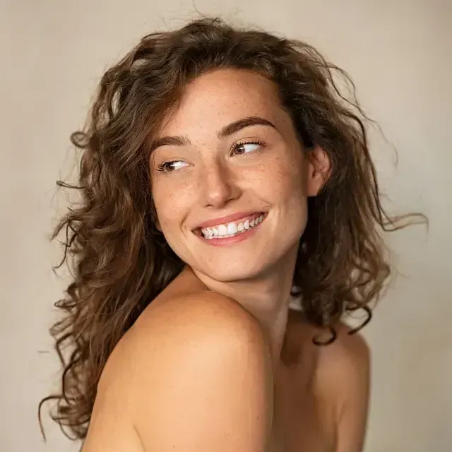 Smiling woman with curly brown hair looking over her shoulder on a neutral background
