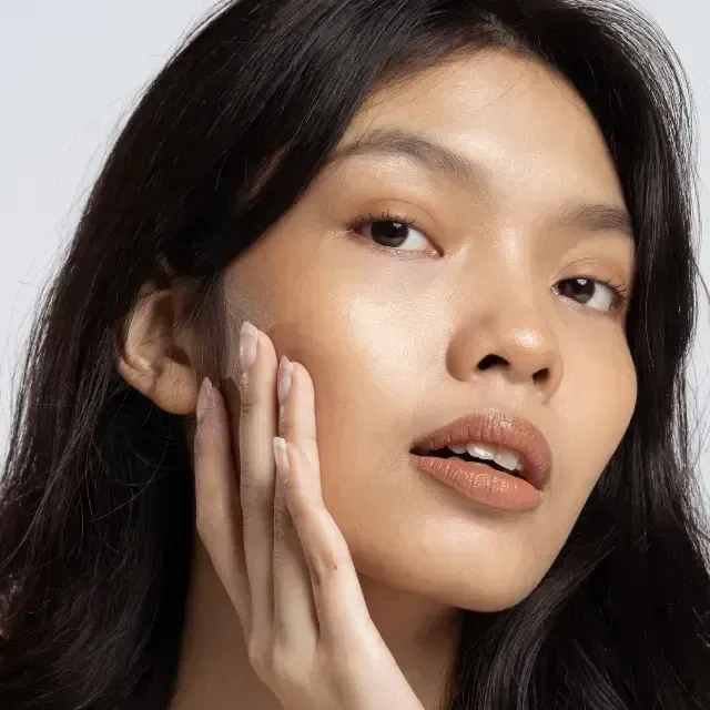 Close-up portrait of a woman with dark hair, resting her hand on her cheek against a light background