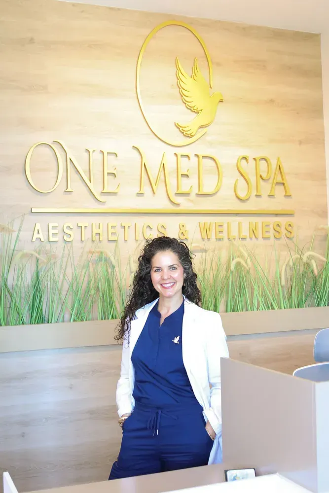 Woman standing in front of One Med Spa reception sign, wearing a white coat and navy scrubs