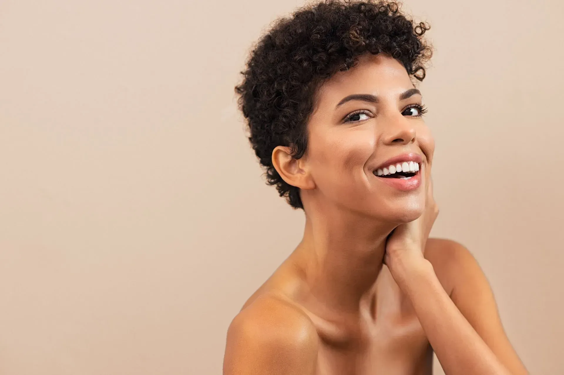Smiling person with short curly hair, touching neck against a beige background