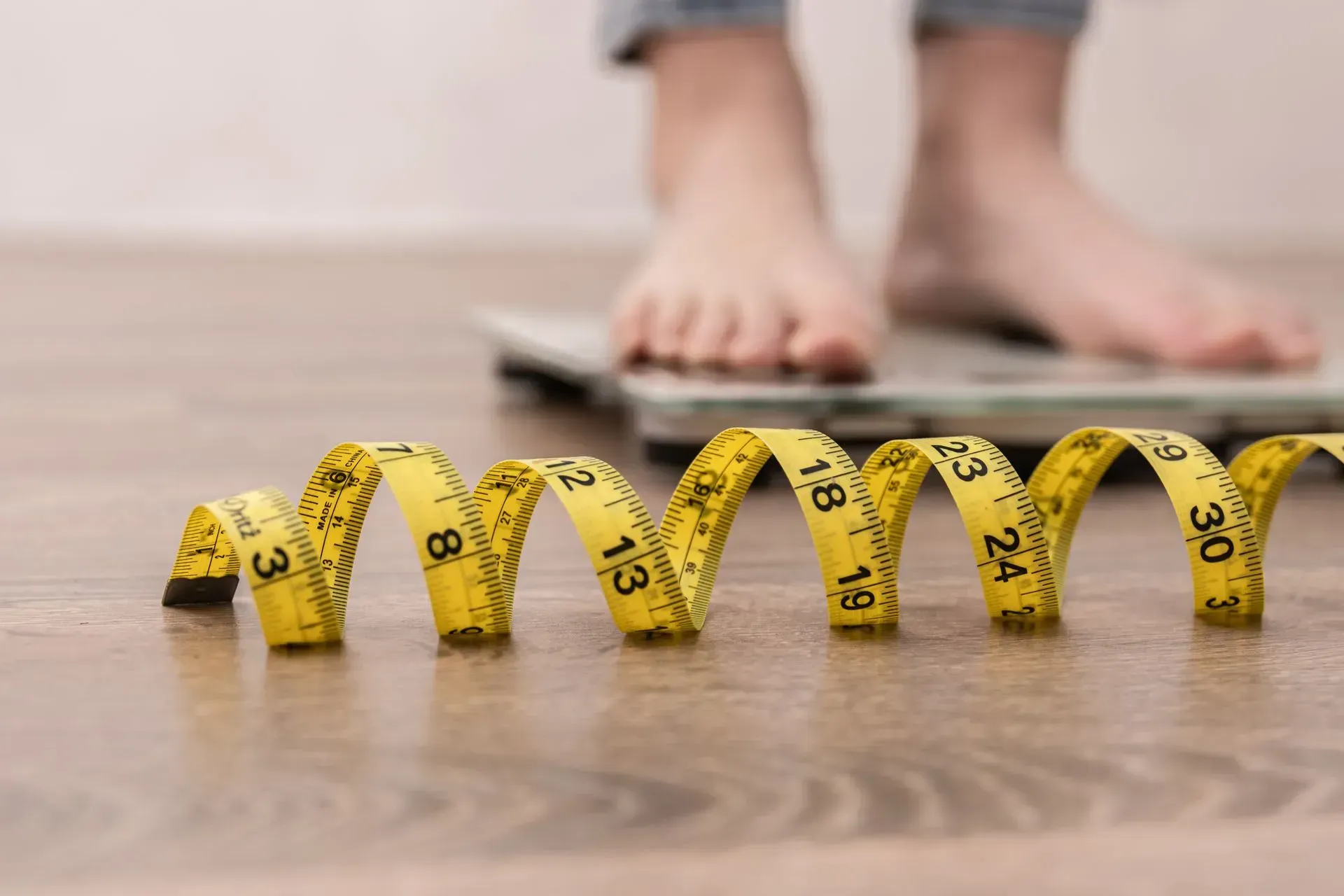 Yellow measuring tape on floor in front of a person standing on a scale, suggesting weight measurement