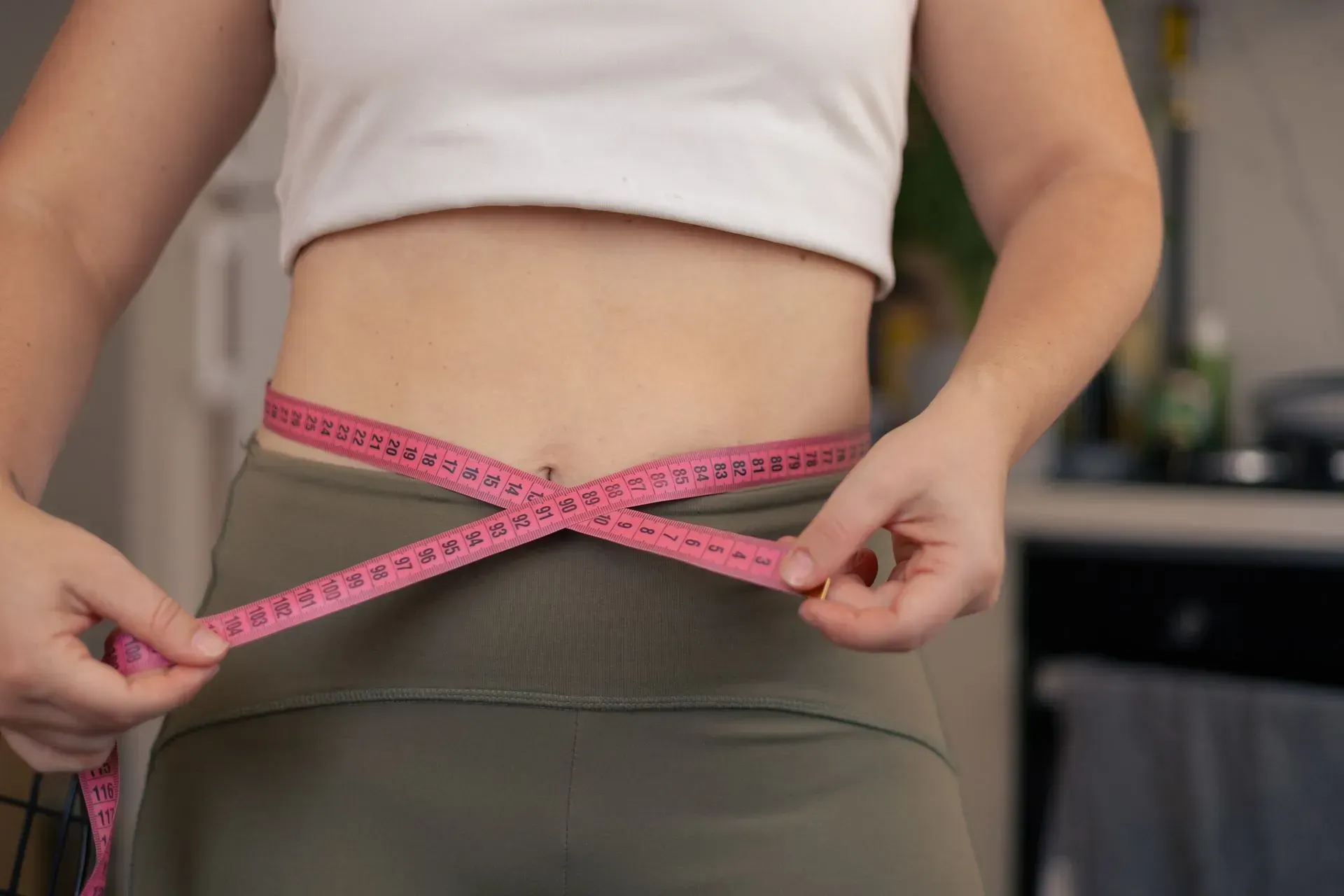 Person measuring waist with pink tape measure over a white top and olive pants.