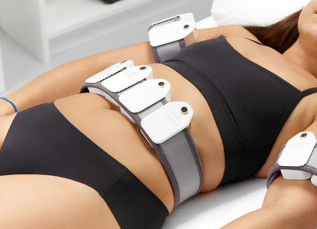 Woman in black sportswear using a gray abdominal muscle stimulation belt on a bed