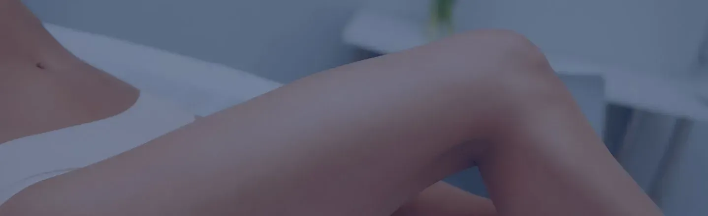 Close-up of a person’s bare legs resting on a white bed with a soft blue-tinted background