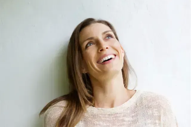 Woman smiling and looking up beside a light gray wall, wearing a cream sweater