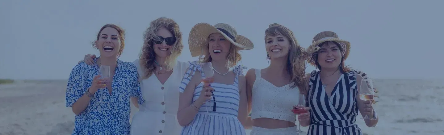 Five friends smiling on a beach, holding drinks in bright summer outfits.