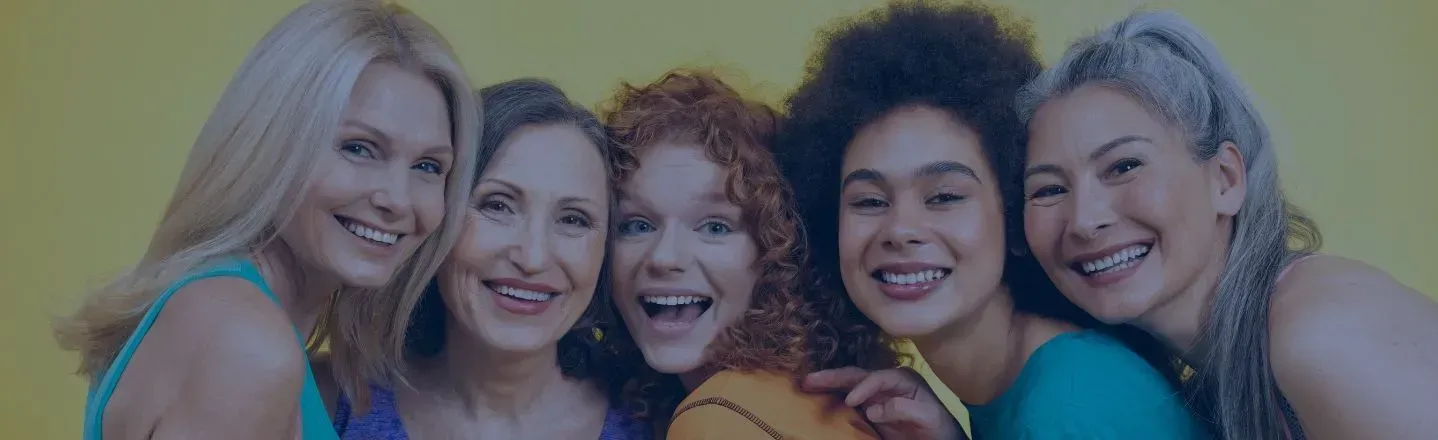 Five smiling women posing close together outdoors against a blurred green background.