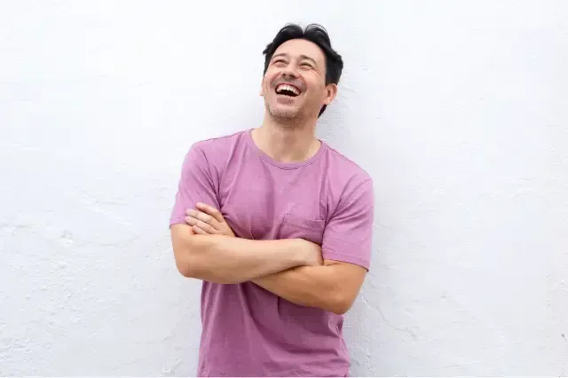 Man in a pink shirt laughing with arms crossed against a white wall