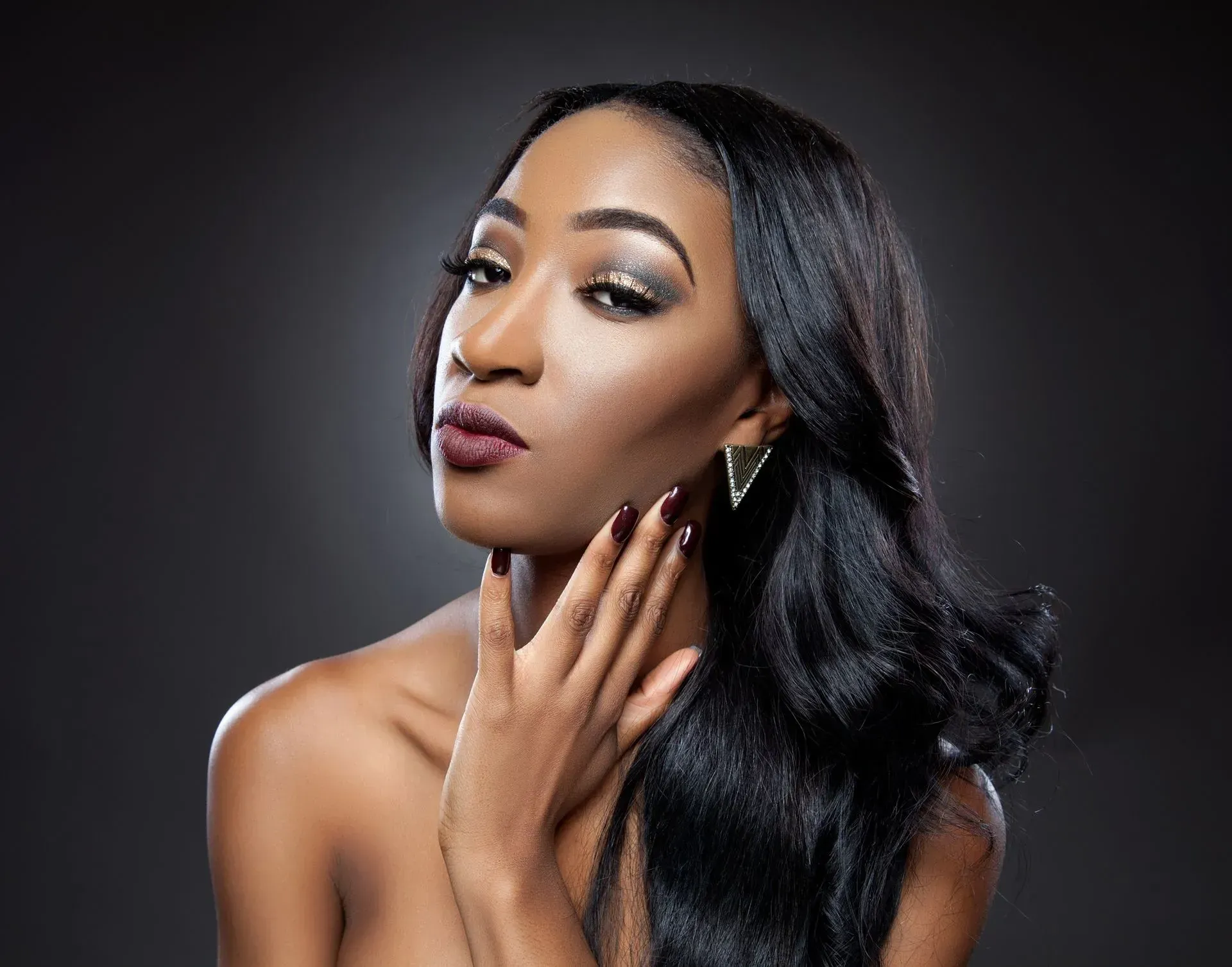 Portrait of a woman with long black hair posing against a dark background, hand on neck, wearing earrings