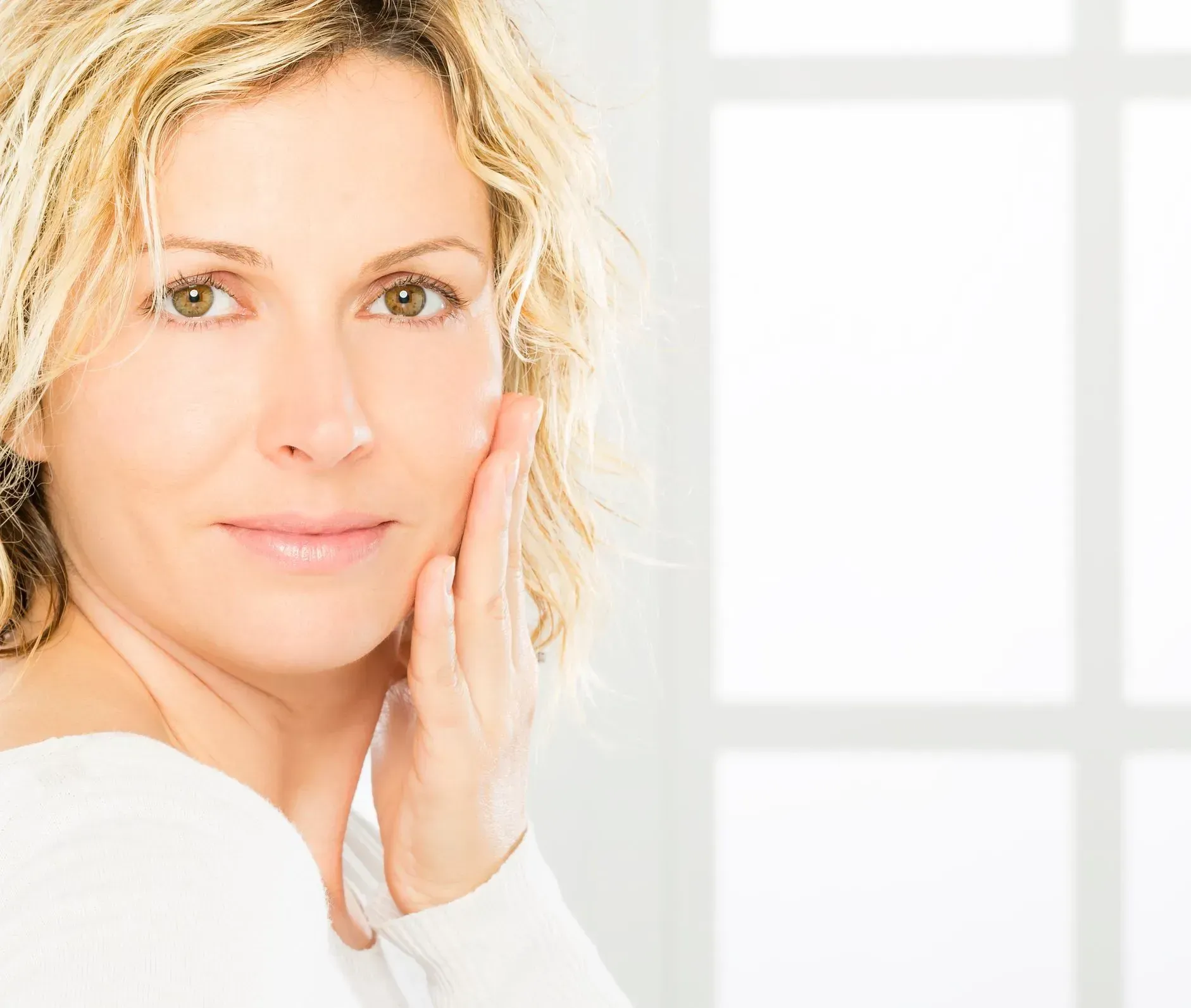 Close-up of a smiling person with hand on cheek against a bright window background