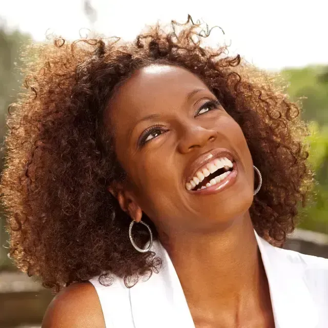 Smiling woman with curly hair looking upward outdoors in a white top