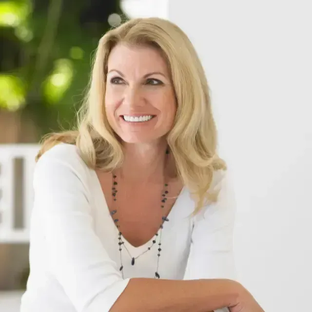 Smiling blonde woman in a white blouse, seated indoors with a bright blurred background