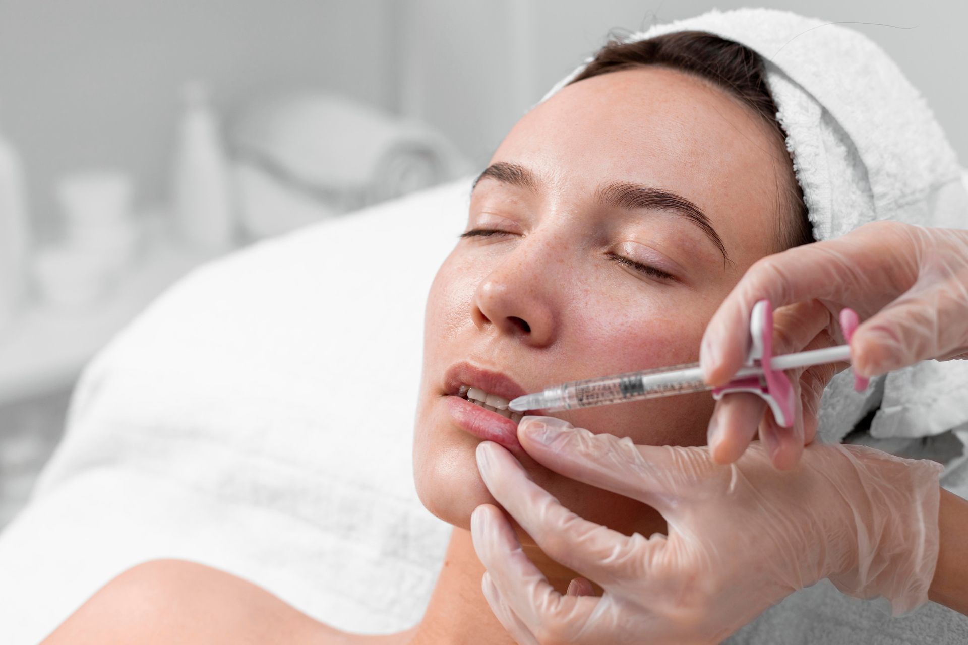 Cosmetic injection to lips by gloved hand while a woman lies with eyes closed on a treatment bed