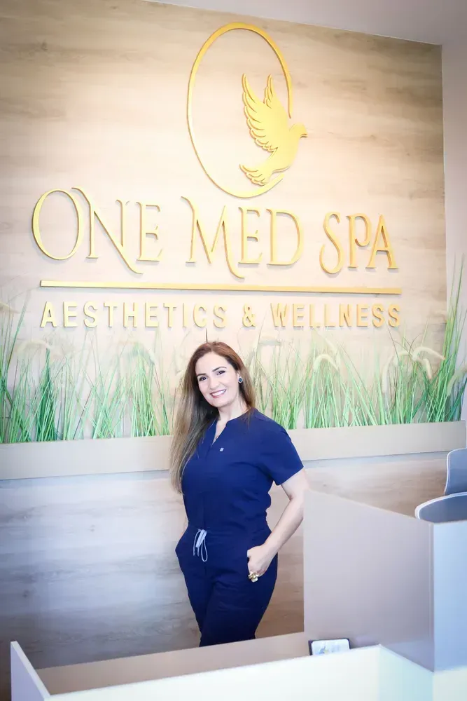 Reception area at One Med Spa with gold logo wall and a woman standing behind the front desk