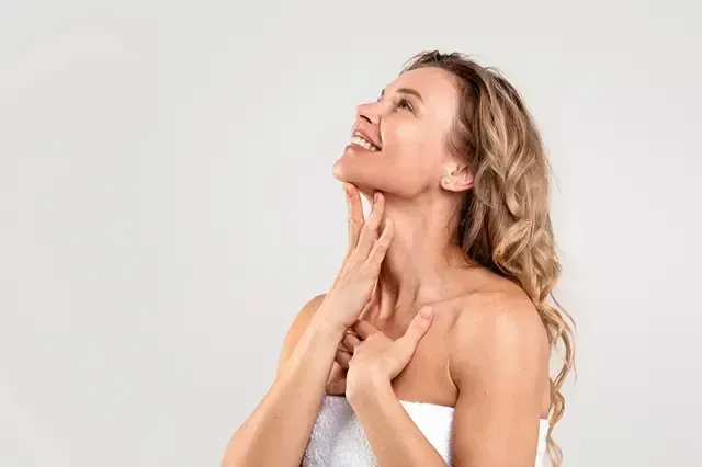 Smiling woman in a white towel applying skincare to her neck against a light background