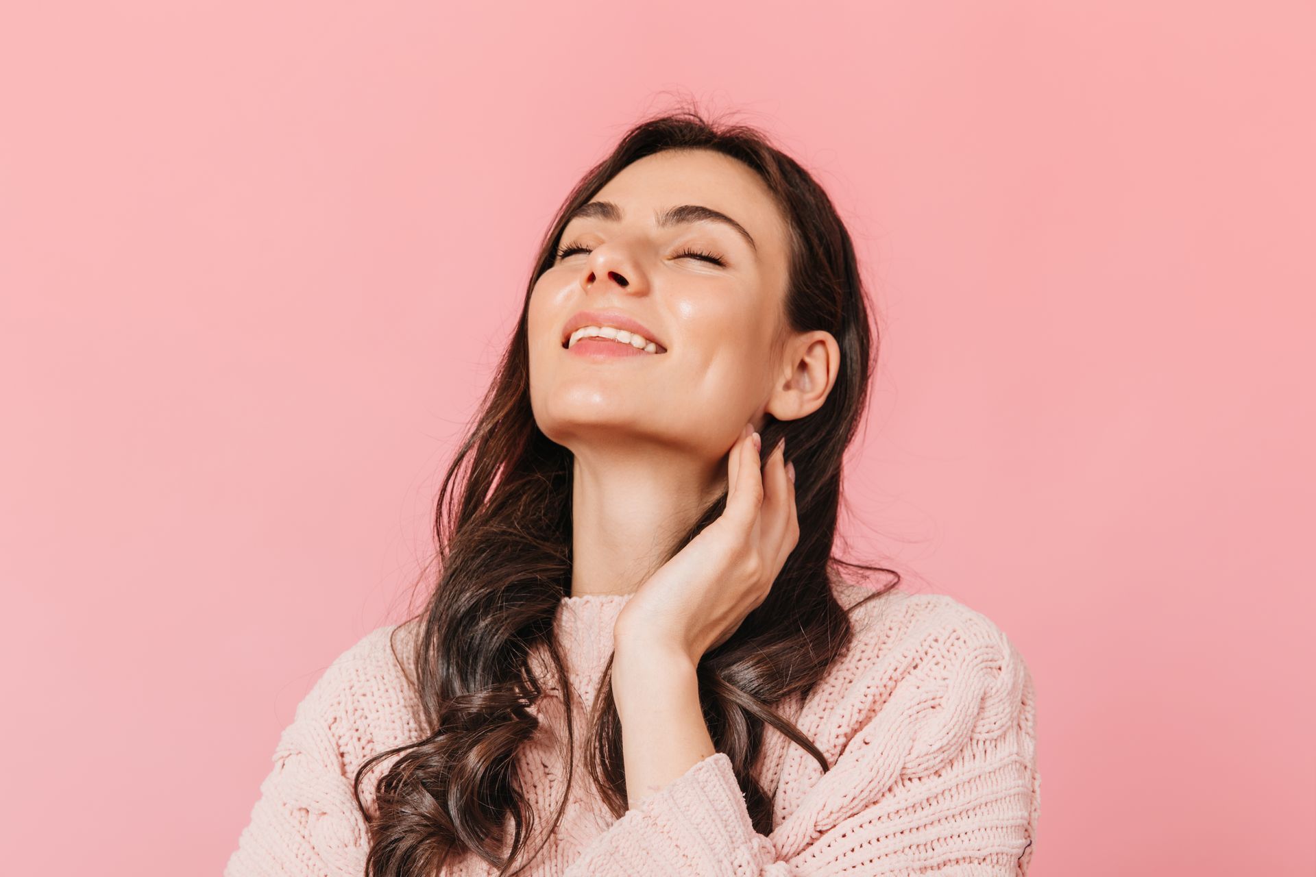Smiling woman with eyes closed, touching her neck, against a pink background