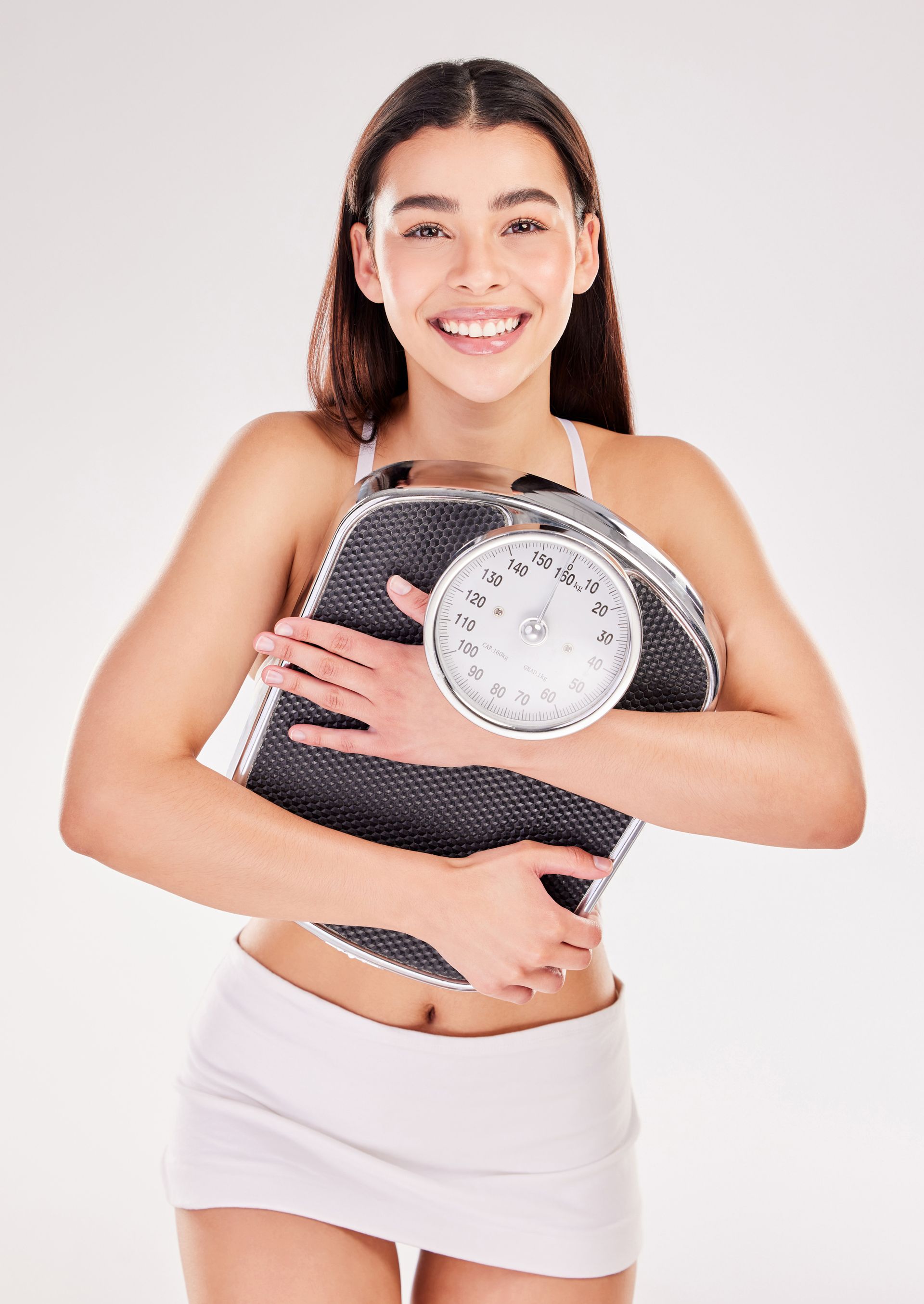 Smiling woman holding a bathroom scale and folded towel against a white background