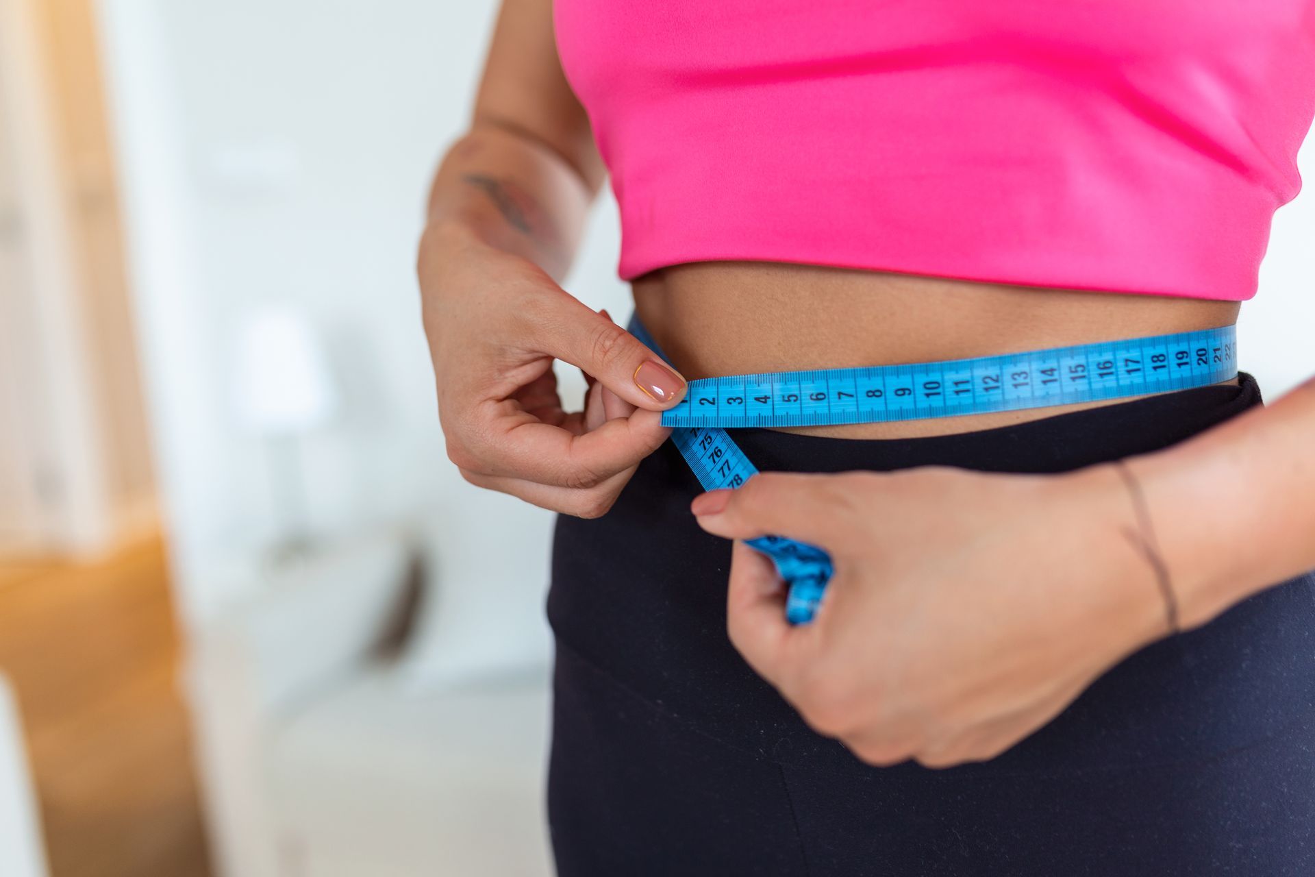 Person measuring waist with blue tape measure over black pants and pink top