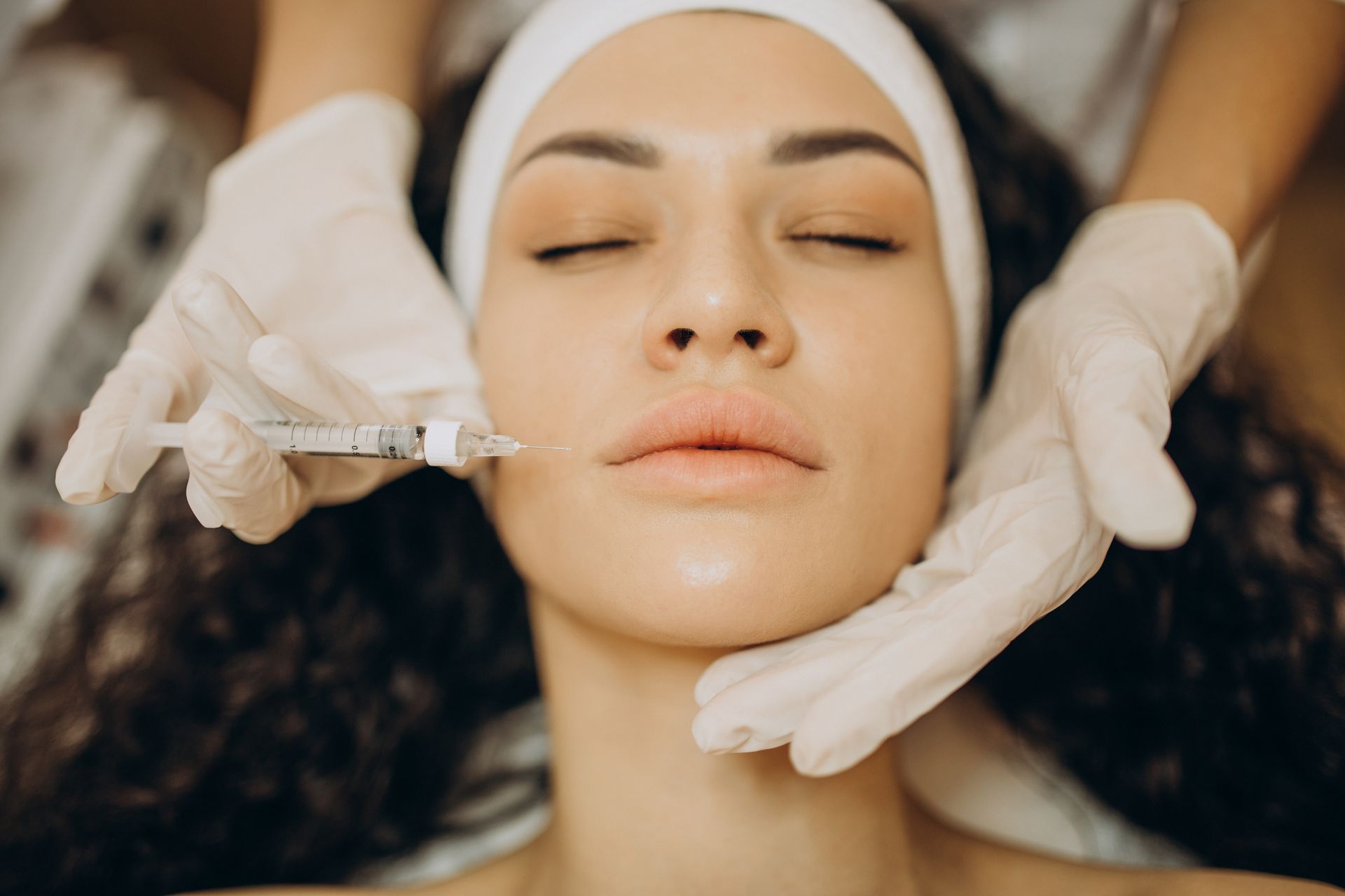 Woman receiving a facial treatment while gloved hands apply syringes near her face