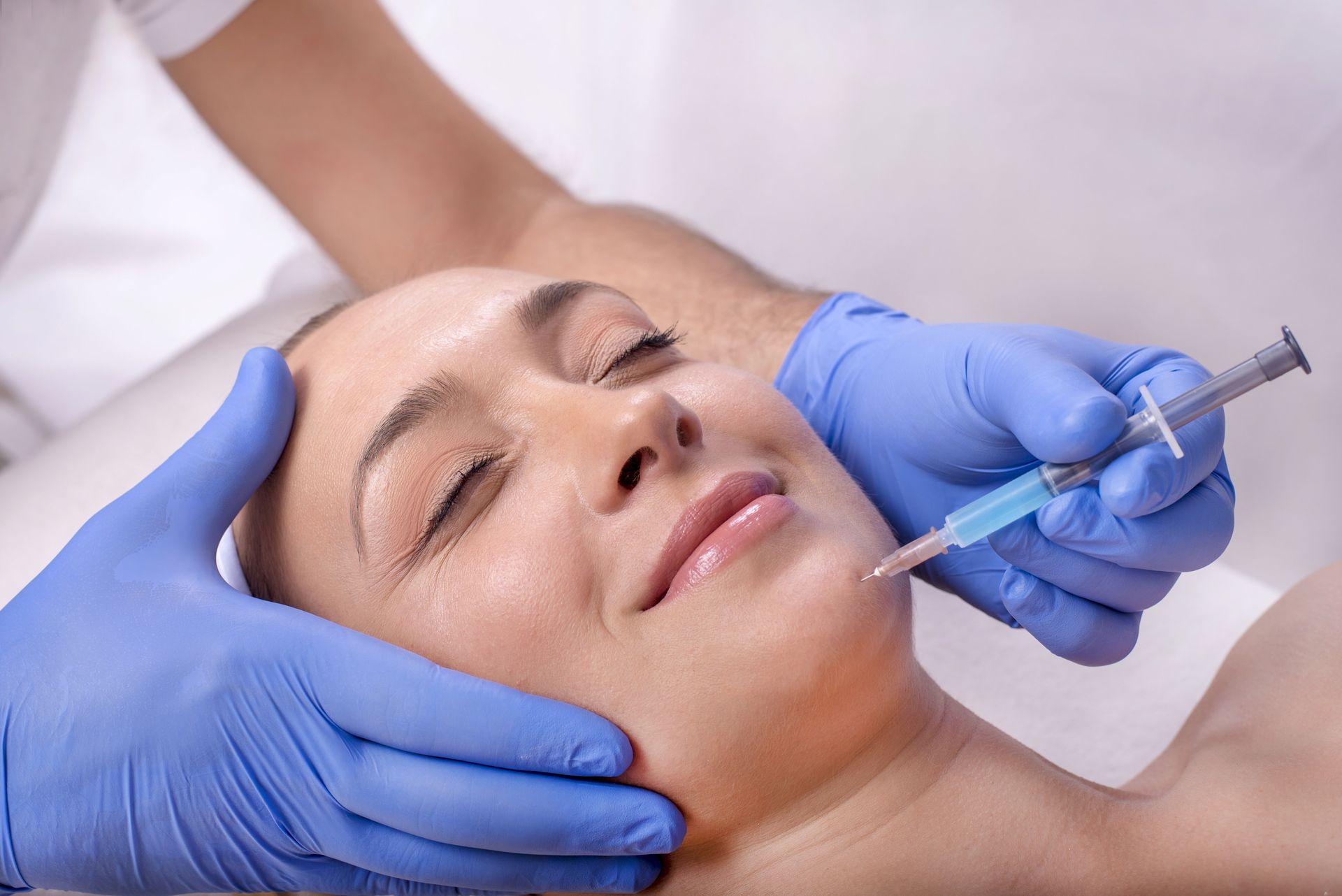 Blue-gloved hands inject a facial treatment into a relaxed woman lying down