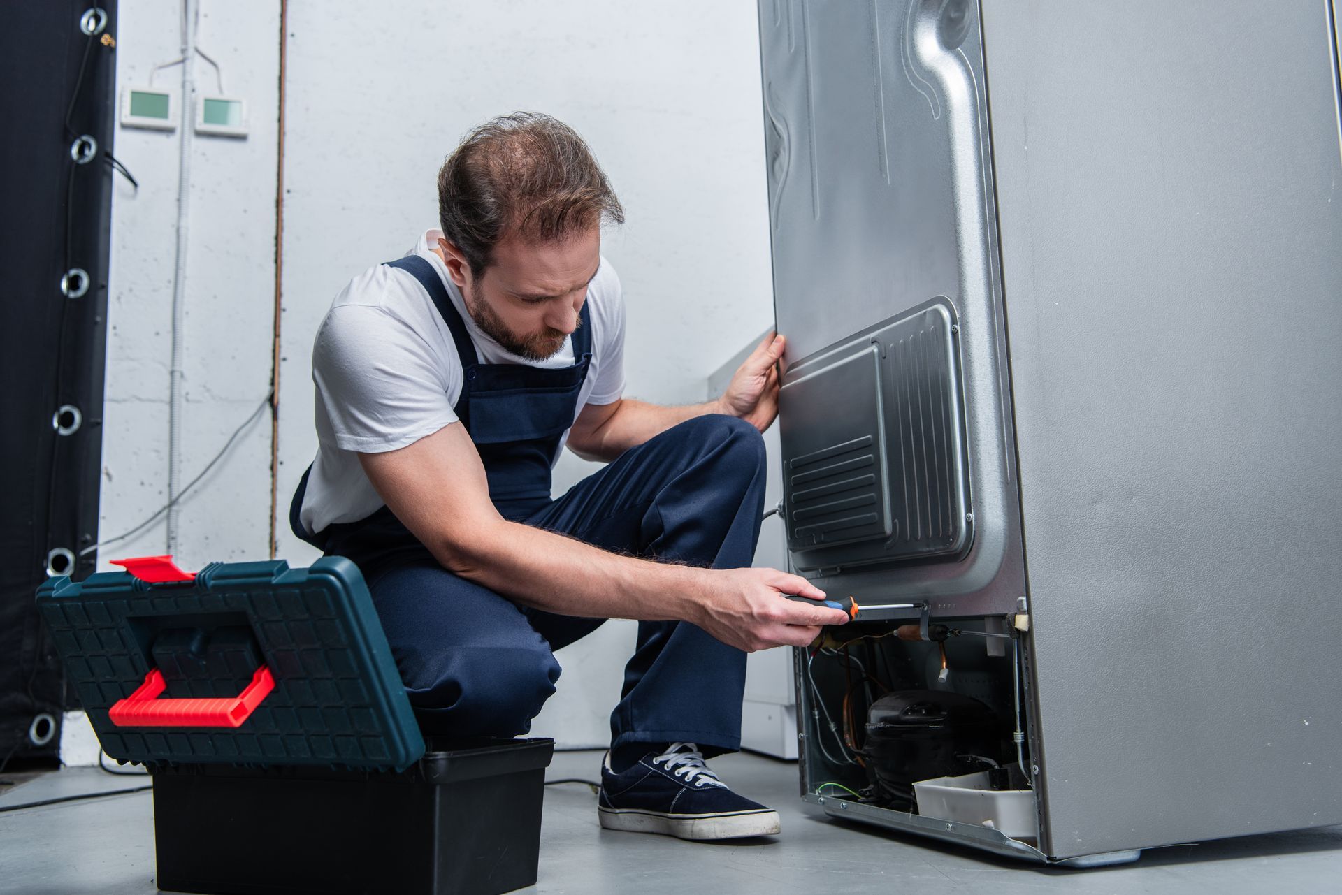 Appliance repair technician inspecting and fixing refrigerator compressor, performing maintenance.