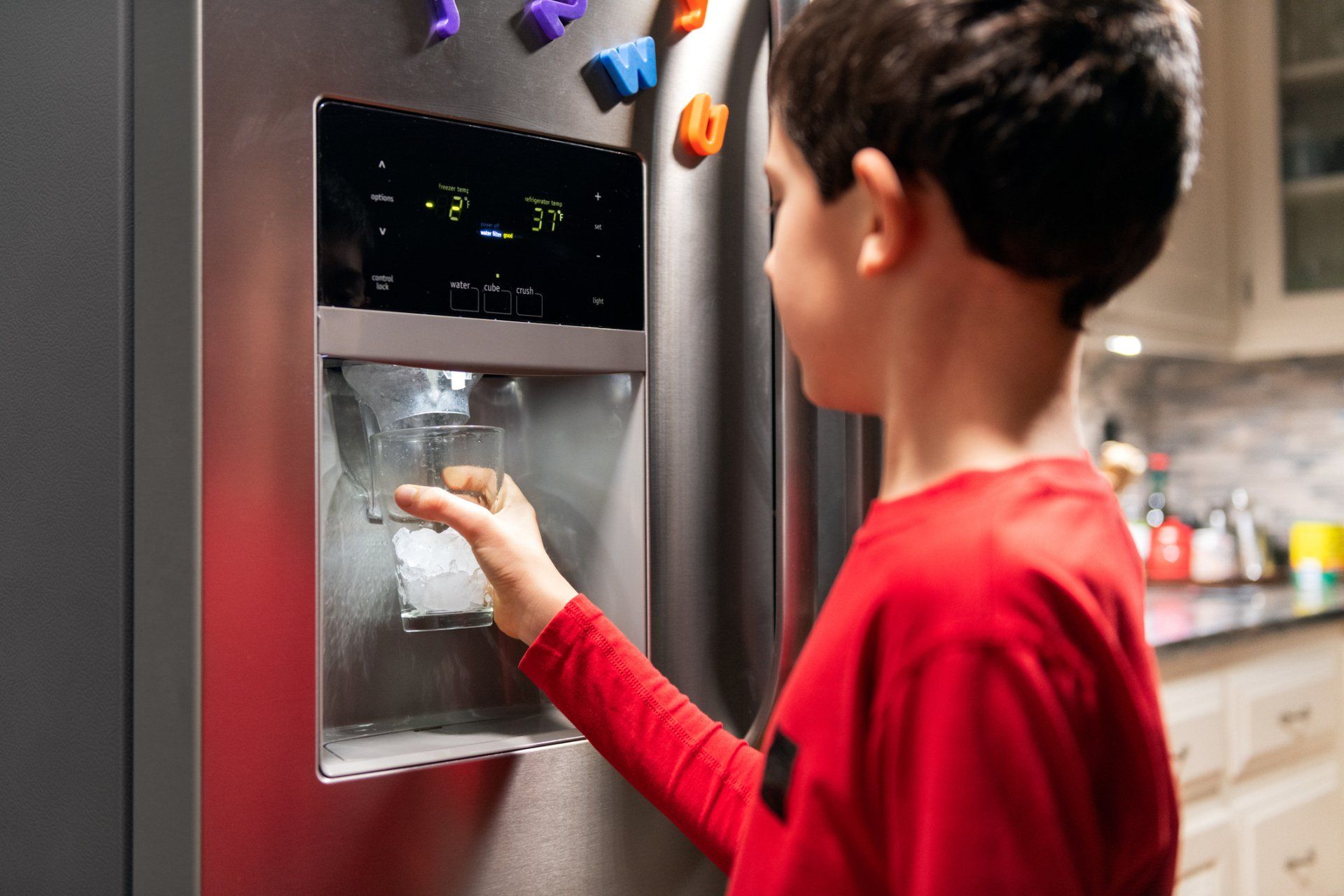 Boy in red shirt getting water from refrigerator dispenser.