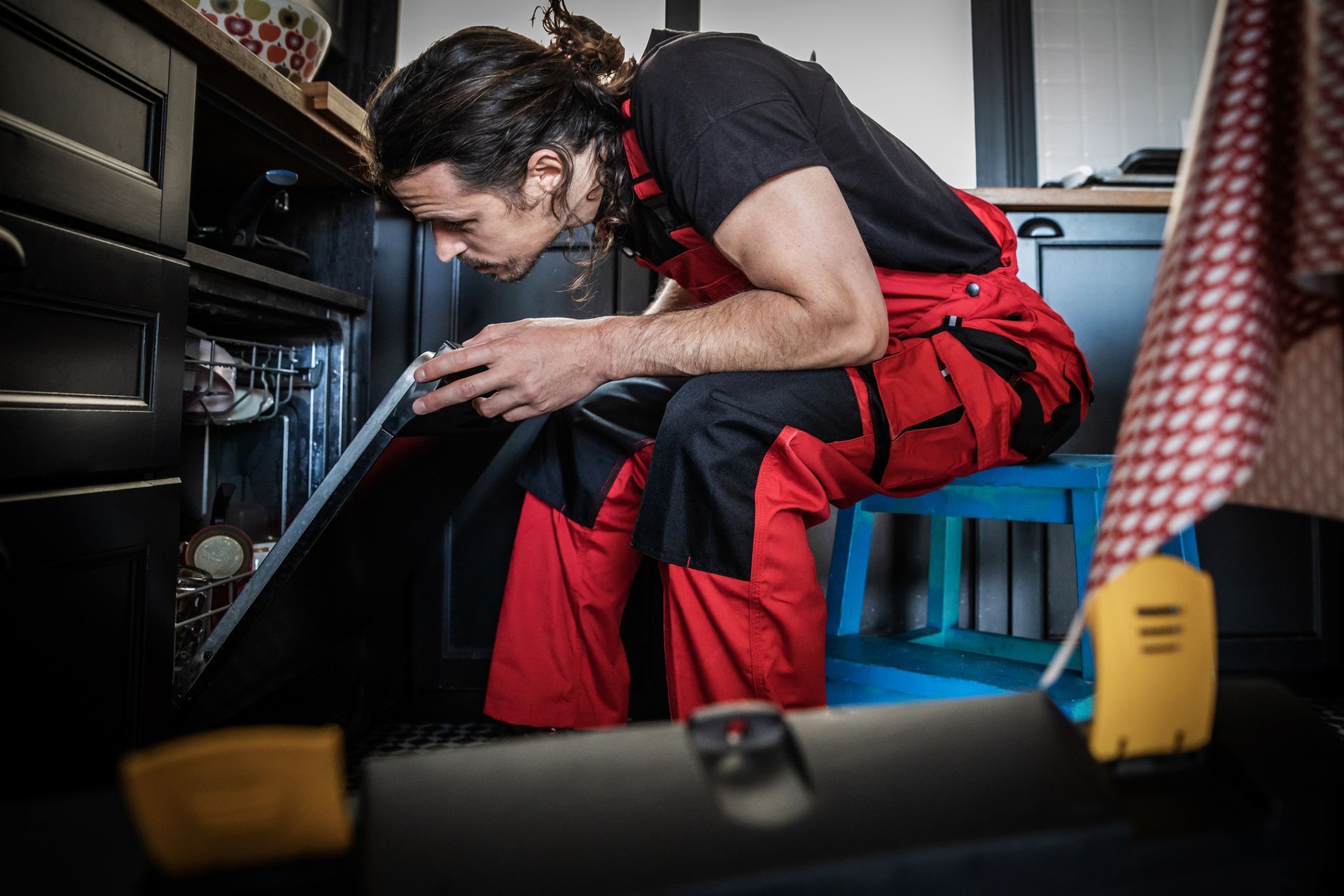 A man in red overalls repairs an oven, sitting on a blue stool in a kitchen.